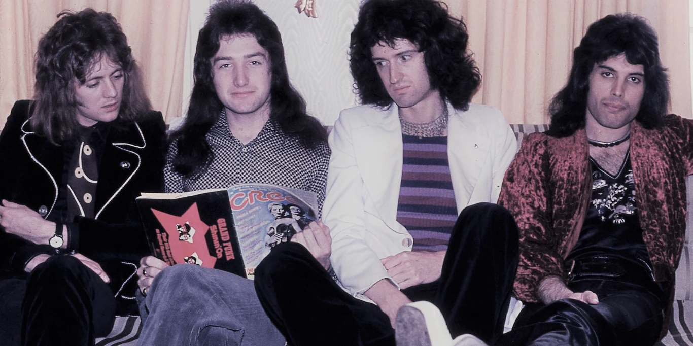 Queen w 1974 r. Czterech mężczyzn siedzących na kanapie, ubrani w wyraziste, stylowe stroje z lat 70., jeden z nich trzyma kolorowy magazyn. W tle jasna zasłona.