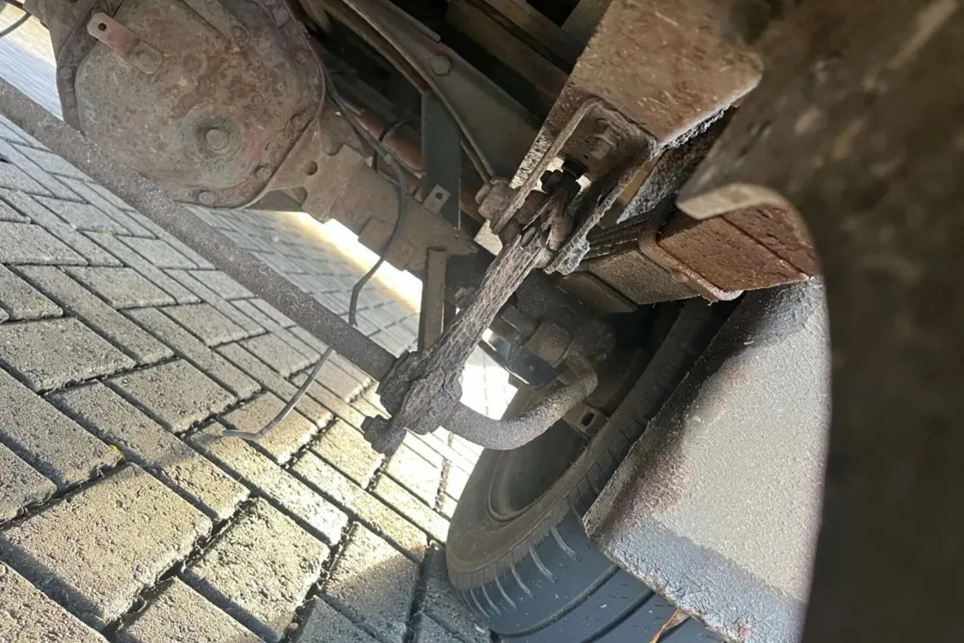 Widok na zawieszenie pojazdu wraz ze sprężynami piórowymi, elementami osi oraz fragmentem koła, z wyraźnie widocznymi oznakami korozji i zużycia mechanicznego.