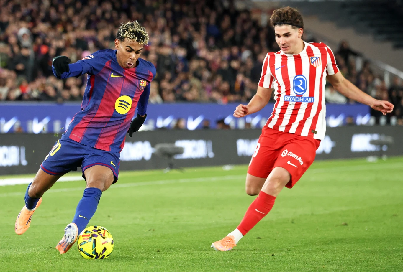 Dwóch piłkarzy walczy o piłkę na boisku podczas meczu, zawodnik w stroju FC Barcelony próbuje dryblować, a piłkarz Atletico Madryt stara się przejąć piłkę, na trybunach widoczna publiczność.