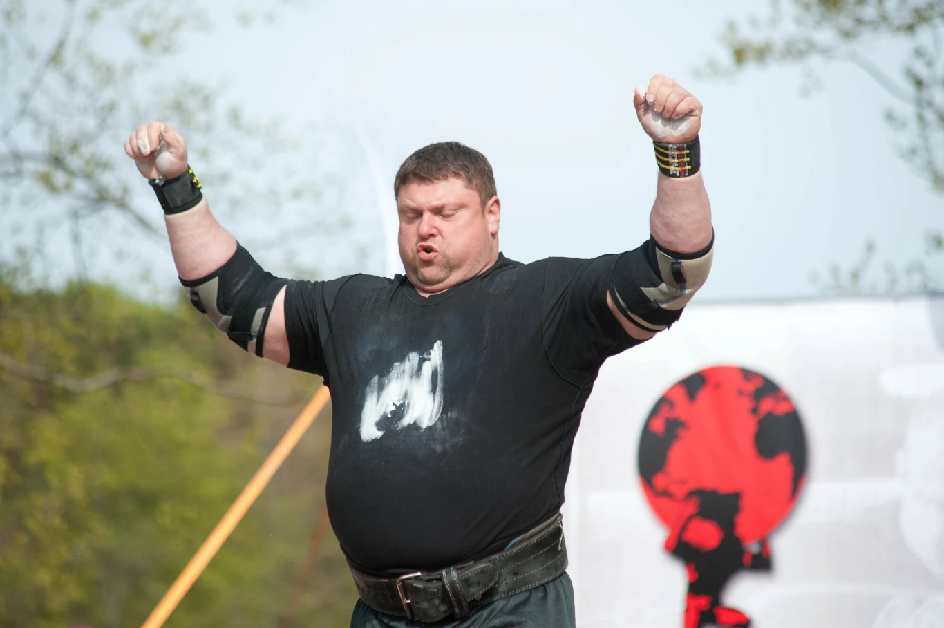 Zydrunas Savickas zdetronizował Mariusza Pudzianowskiego w 2009 roku Mężczyzna o silnej posturze z podniesionymi uniesionymi ramionami, ubrany w czarną koszulkę z widocznym białym śladem kredy na piersi oraz ochraniaczami na nadgarstkach i łokciach. W tle rozmyty baner z grafiką globu.