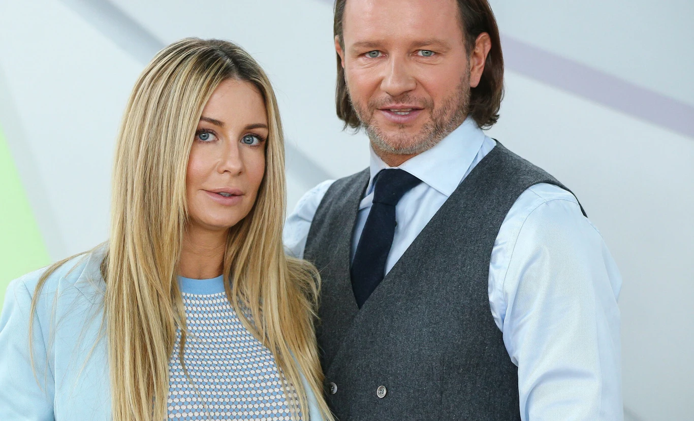 Para kobiet i mężczyzna w eleganckiej stylizacji, kobieta ma długie blond włosy i jasną marynarkę, mężczyzna ubrany jest w szary kamizelkę i białą koszulę z krawatem, stoją blisko siebie na jasnym tle.