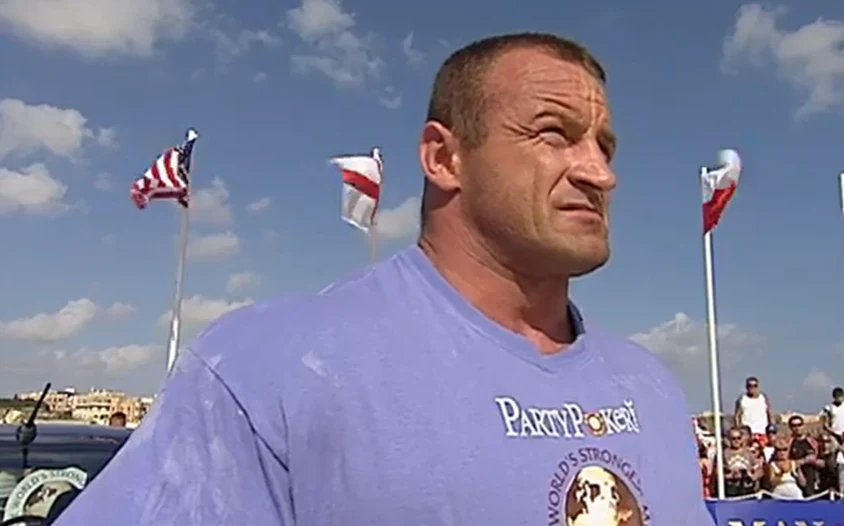 Tak wyglądał ostatni taniec Pudzianowskiego Silnie zbudowany mężczyzna ubrany w jasnofioletową koszulkę z napisem oraz grafiką, stojący na tle flag i nieba, wokół grupy ludzi obserwujących wydarzenie.