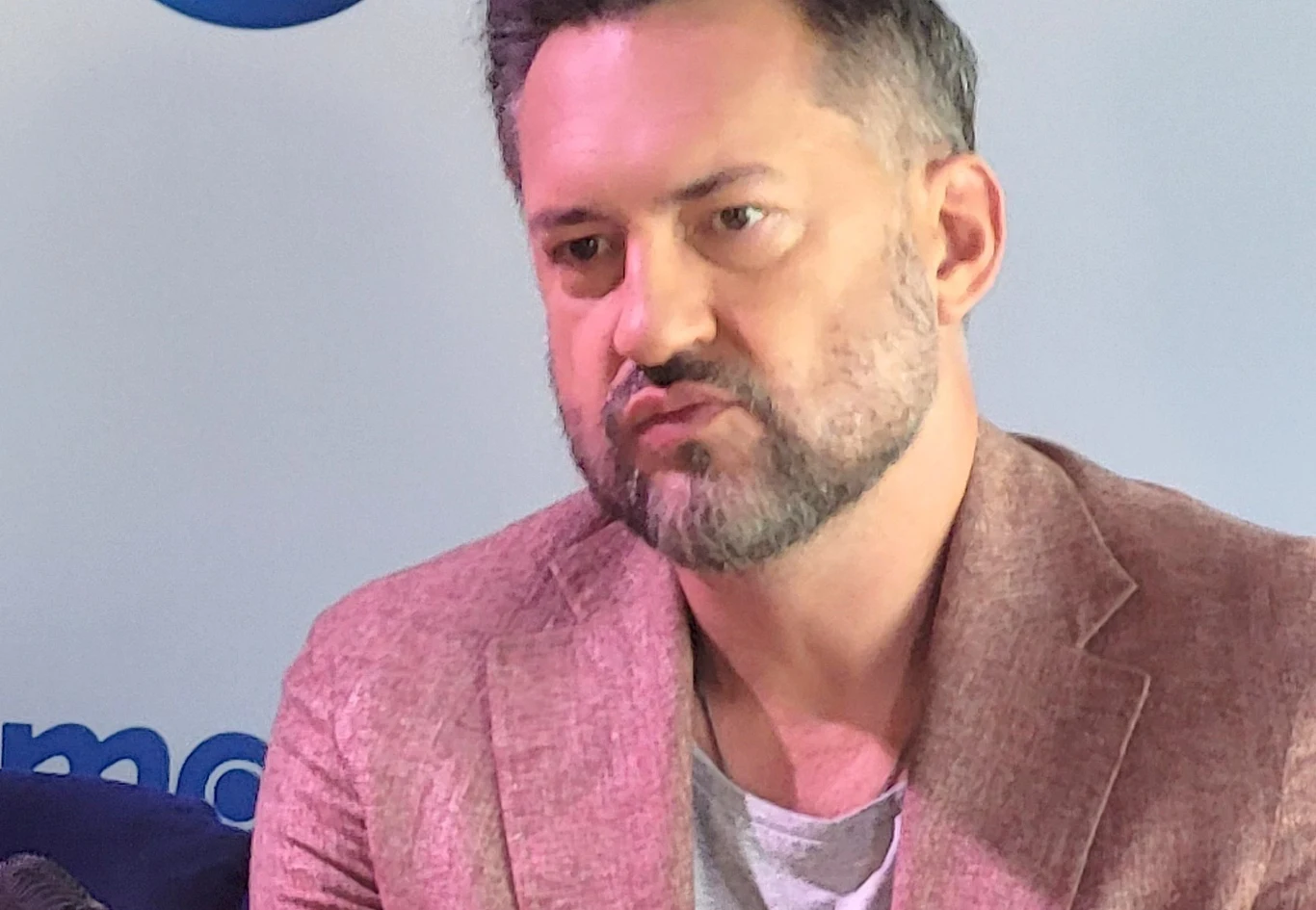 Mężczyzna z brodą i krótkimi ciemnymi włosami ubrany w jasnobrązową marynarkę i szary T-shirt, siedzi na tle jasnej ściany z niebieskimi elementami graficznymi. Jego wyraz twarzy jest poważny i skupiony.