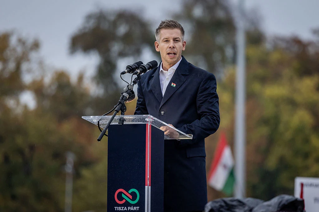 Péter Magyar, lider TISZA i główny przeciwnik Viktora Orbána Mężczyzna ubrany w elegancki płaszcz wygłasza przemówienie stojąc przy mównicy z mikrofonami, na której widnieje logo Tisza Párt, w tle jesienne drzewa i zamazana flaga narodowa.