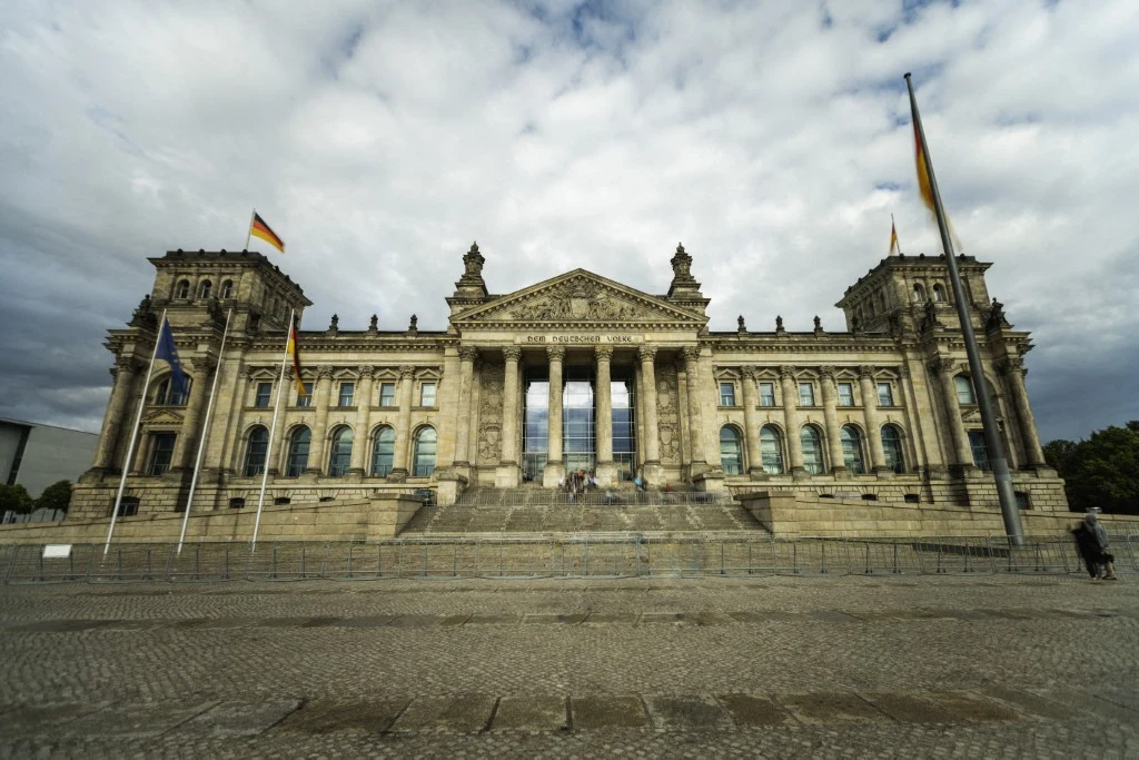 Niemcy wezwali rosyjskiego ambasadora. W tle poważne oskarżenia. Na zdj. budynek Reichstagu Monumentalna fasada gmachu parlamentu niemieckiego Reichstag z wysokimi kolumnami i schodami wejściowymi, ozdobiona flagami Niemiec i Unii Europejskiej, w tle zachmurzone niebo.