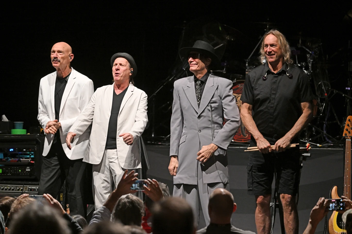 Pod szyldem Beat występują Tony Levin, Adrian Belew, Steve Vai i Danny Carey Czterech mężczyzn stoi na scenie koncertowej przed publicznością, trzech ubranych w jasne garnitury, jeden w czarną koszulę i krótkie spodenki, jeden z muzyków trzyma pałeczki do perkusji, za nimi widoczne są bębny i inny sprzęt muzyczny.