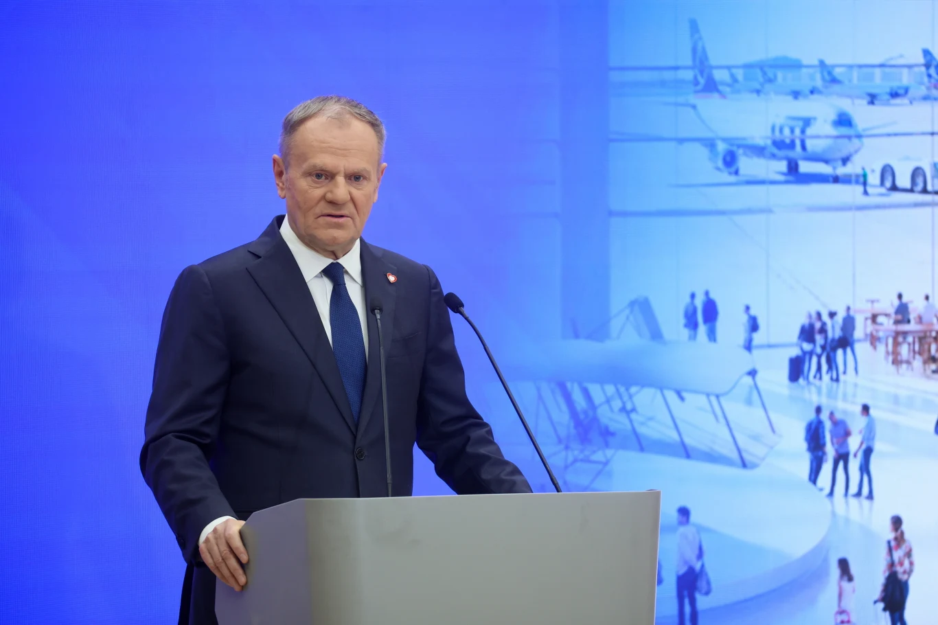 Port Polska. Premier Donald Tusk ogłosił nową nazwę portu lotniczego Mężczyzna w garniturze przemawia przy mównicy na tle niebieskiej grafiki z motywami lotniczymi, w tym rysunkami samolotów i ludzi w hali lotniska.