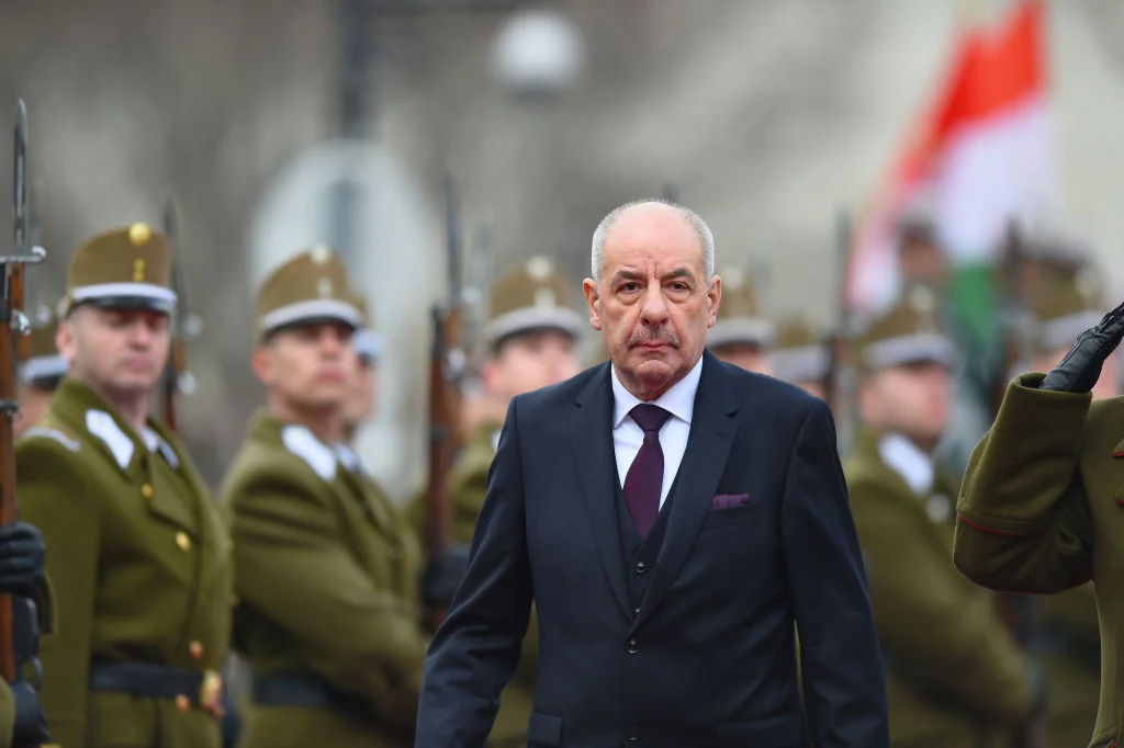 Prezydent Węgier Tamás Sulyok Starszy mężczyzna w garniturze idzie przed szeregiem umundurowanych żołnierzy trzymających broń z bagnetami. W tle widać rozmazaną flagę narodową. Atmosfera oficjalna, podniosła.