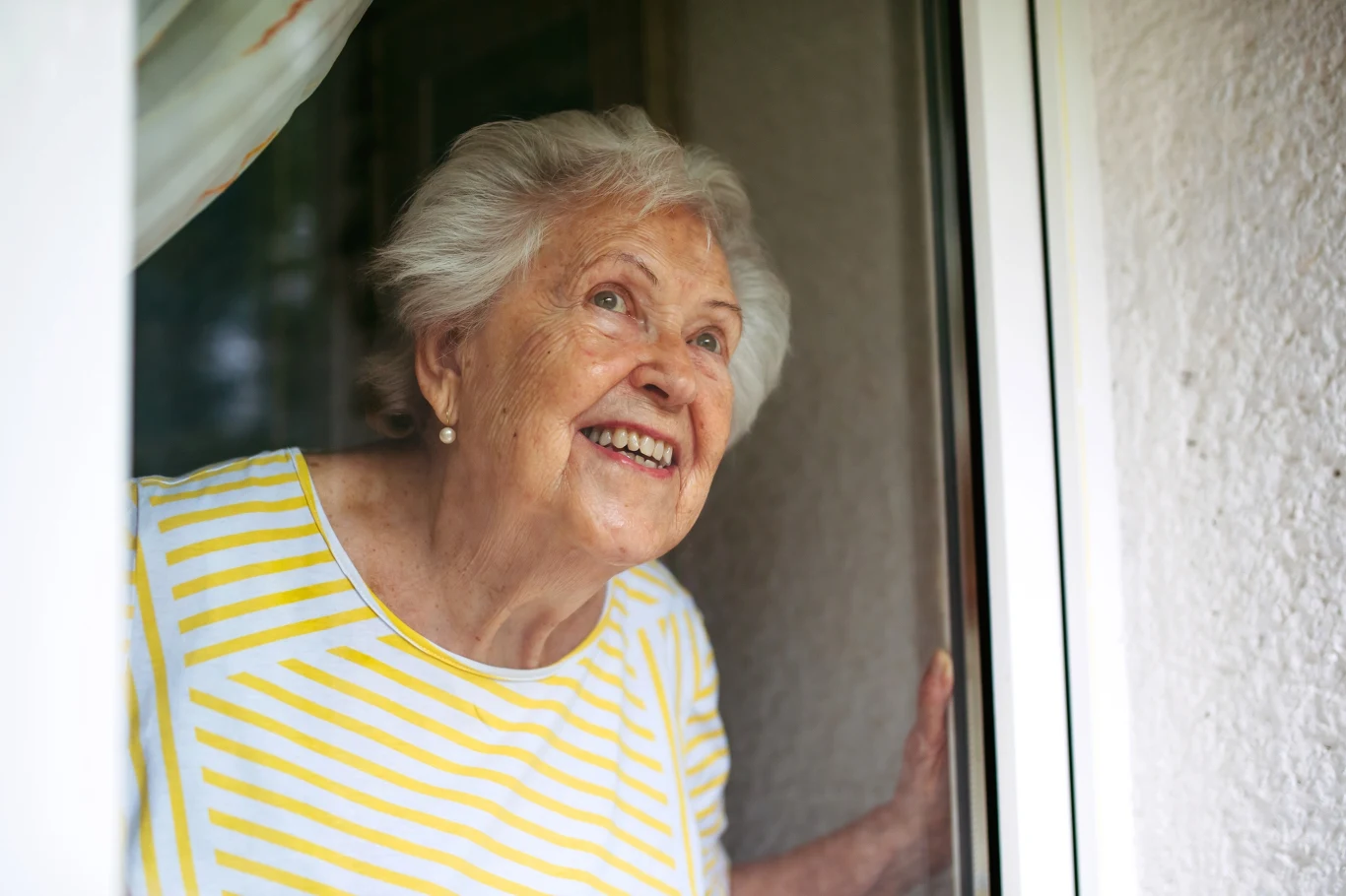 Starsza kobieta w białej bluzce w żółte paski stoi w otwartym oknie, uśmiecha się szeroko i spogląda na zewnątrz. Jej twarz wyraża radość i zaciekawienie, a za nią widoczna jest biała zasłona i ściana.