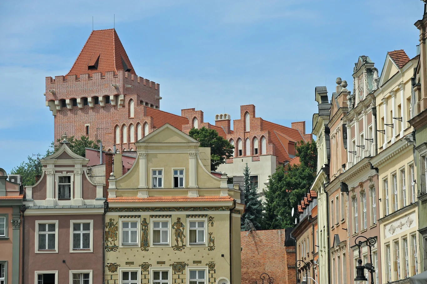 Ceglana wieża zamkowa z czerwonym dachem górująca nad zabytkowymi kamienicami Starego Miasta, ozdobionymi dekoracyjnymi fasadami i detalami architektonicznymi.