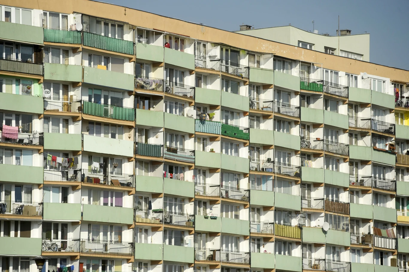 Wielokondygnacyjny blok mieszkalny z licznymi balkonami, na których znajdują się różne przedmioty codziennego użytku, pranie i dekoracje. Fasada budynku ma zielone i beżowe elementy w układzie poziomym.