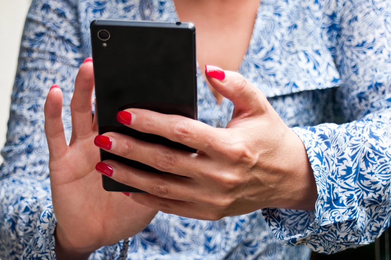 Dłonie kobiety z czerwonym lakierem na paznokciach trzymające czarny smartfon, ubrana w niebieską koszulę z białym roślinnym wzorem.