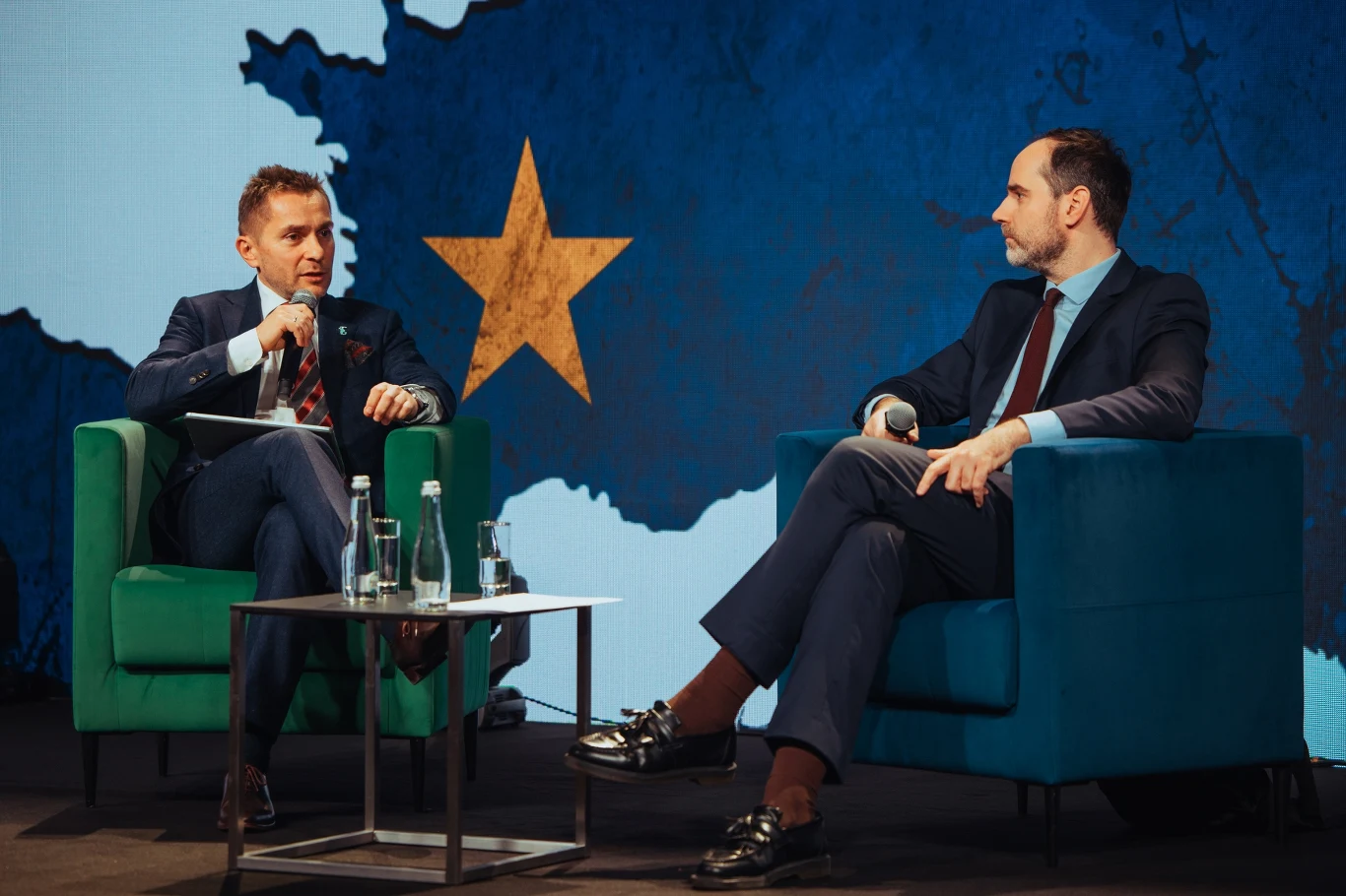 Dwóch mężczyzn w garniturach siedzi na scenie w wygodnych fotelach, prowadząc rozmowę na tle dużej ilustracji z mapą Europy i żółtą gwiazdą, na stoliku znajdują się butelki z wodą i szklanki.