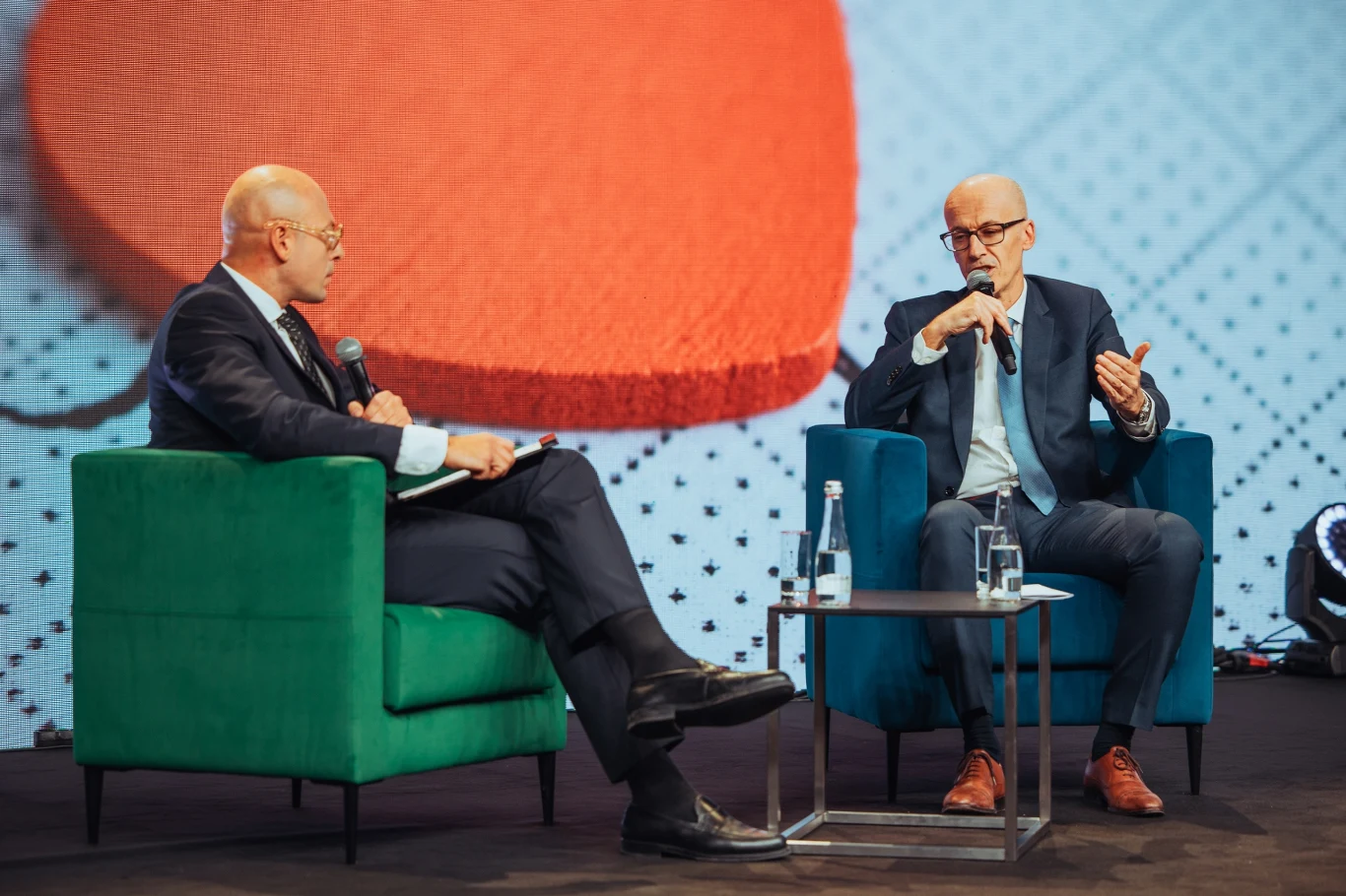 Dwóch mężczyzn w garniturach siedzi naprzeciw siebie na kolorowych fotelach, prowadząc rozmowę z mikrofonami w dłoniach. W tle duży ekran z graficznym wzorem, na stoliku znajdują się butelki z wodą i szklanki.