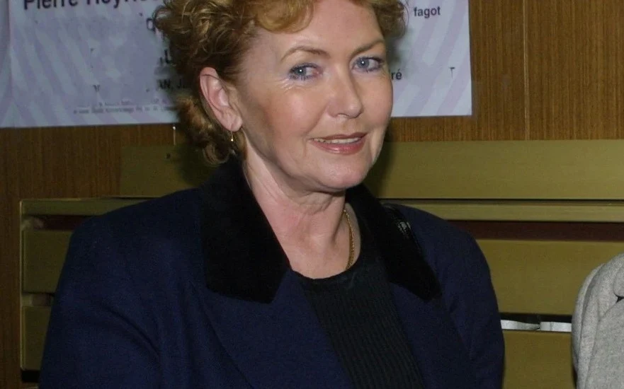 Anna Wanda Głębocka Kobieta o rudych, kręconych włosach w ciemnej marynarce siedzi na tle drewnianych paneli z zamieszczonym na ścianie plakatem. Uśmiecha się lekko i patrzy w stronę obiektywu.