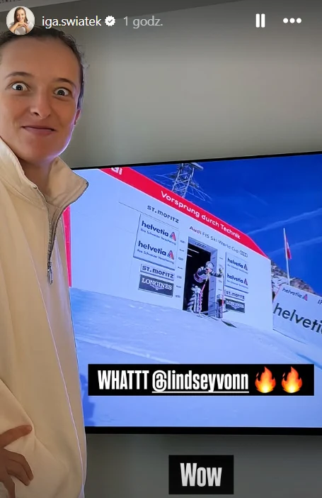 Tak Iga Świątek zareagowała na zwycięstwo Lindsey Vonn Kobieta w jasnej bluzie stoi przed telewizorem, na którym widać scenę sportową z zawodów narciarskich, trybuny i logo sponsorów. Wyraża zaskoczenie lub zachwyt.