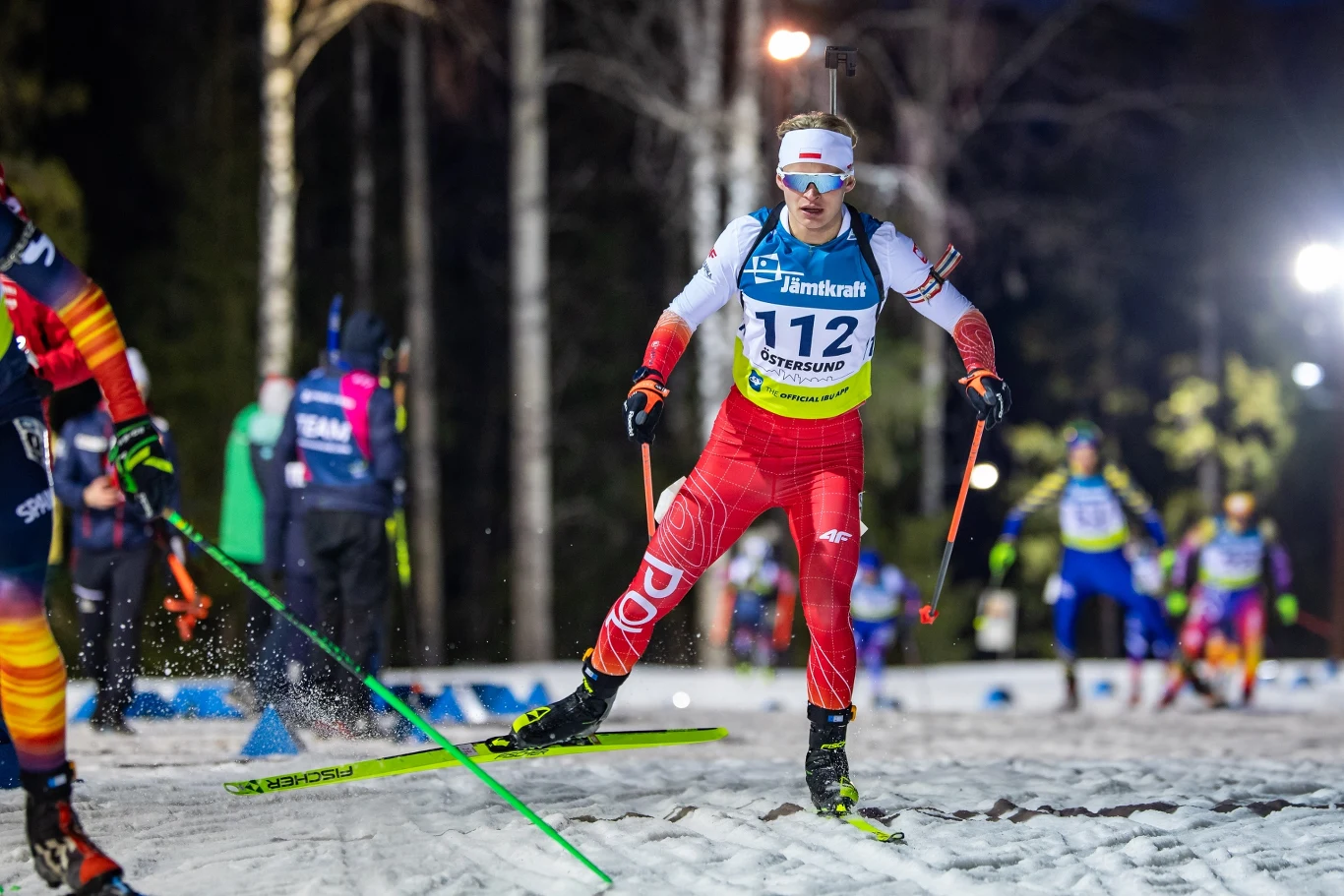 Zawodniczka w czerwonym stroju z numerem 112 dynamicznie jedzie na nartach biegowych po oświetlonej trasie, w tle pozostali uczestnicy wyścigu oraz zimowy, leśny krajobraz.