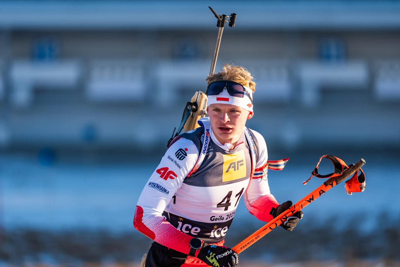 Zawodnik biathlonowy w biało-czerwonym stroju sportowym biegnie z nartami i kijami na tle rozmazanego stadionu, na plecach niesie karabin, na głowie ma opaskę, a jego twarz wyraża skupienie.