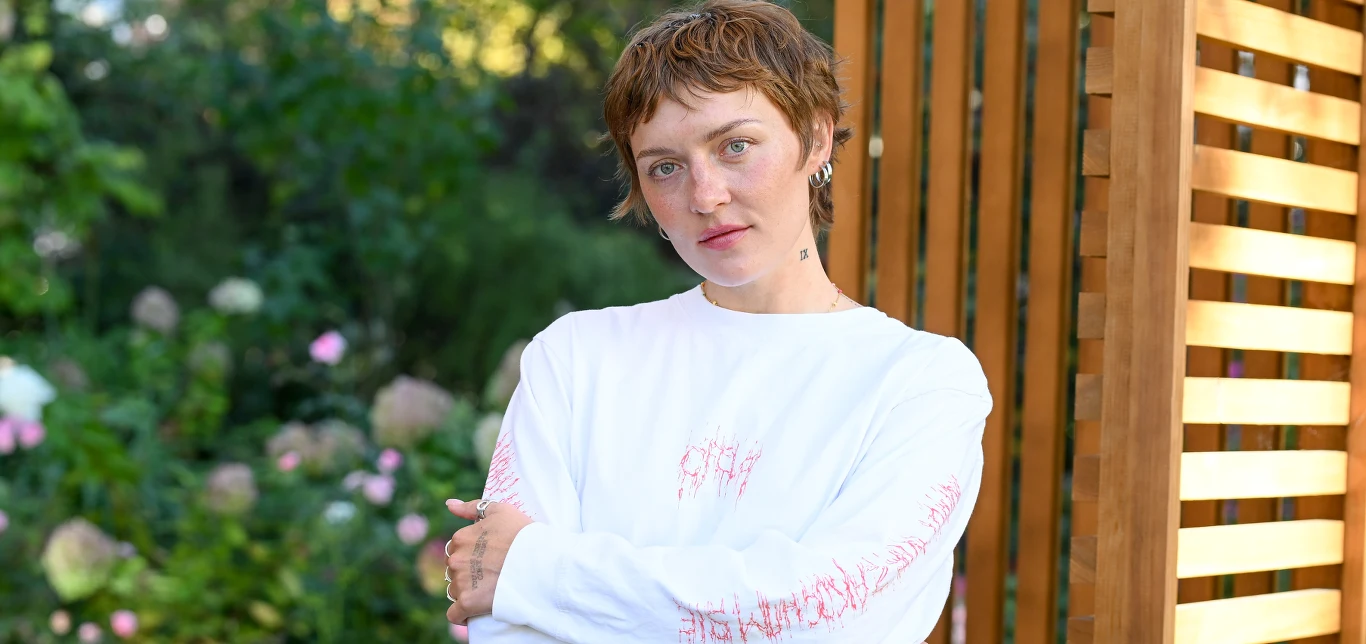 Ofelia Młoda kobieta z krótkimi brązowymi włosami ubrana w biały długi T-shirt stoi na tle ogrodu pełnego kwiatów oraz elementów drewnianej pergoli. Spokojny wyraz twarzy i delikatne światło podkreślają naturalną scenerię.