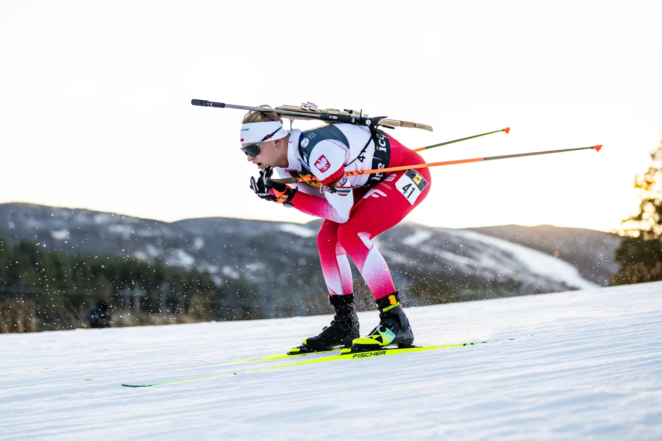 Zawodniczka w kombinezonie z polską flagą i numerem startowym biegnie na nartach biegowych z karabinem na plecach, dynamiczna sylwetka na tle ośnieżonych gór, akcent na szybkość i sportową determinację.