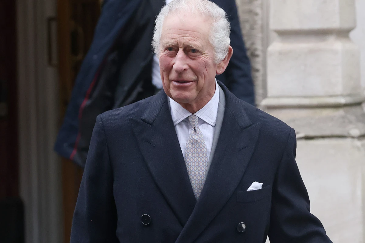 Król Karol III Starszy mężczyzna w eleganckim, ciemnym garniturze i jasnym krawacie stoi na tle jasnej, kamiennej ściany, delikatnie się uśmiecha, w tle widoczne ciemne ubrania.