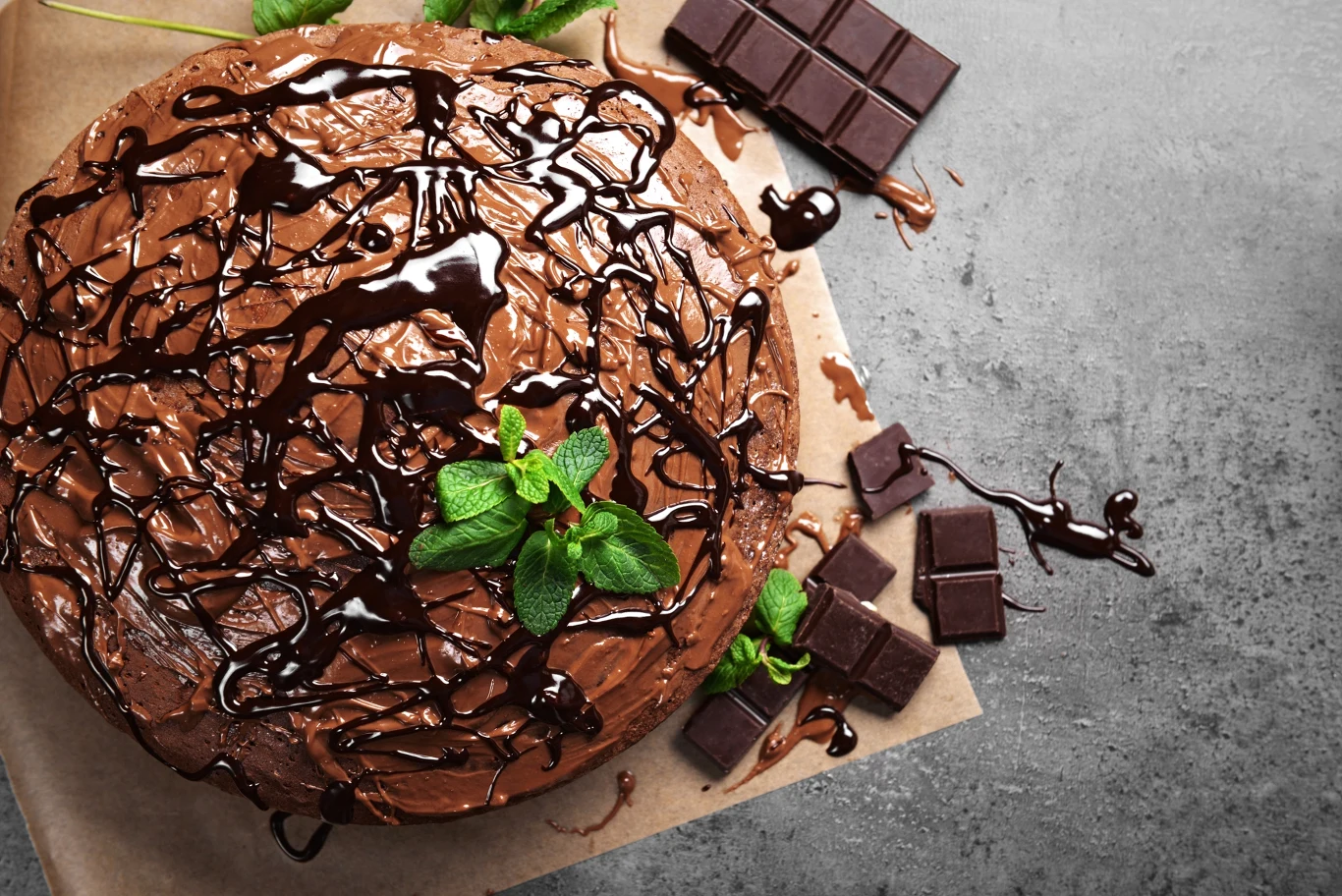 Okrągły tort czekoladowy oblany polewą czekoladową, udekorowany świeżą miętą, obok kawałki gorzkiej czekolady, jasne tło z papierem do pieczenia.