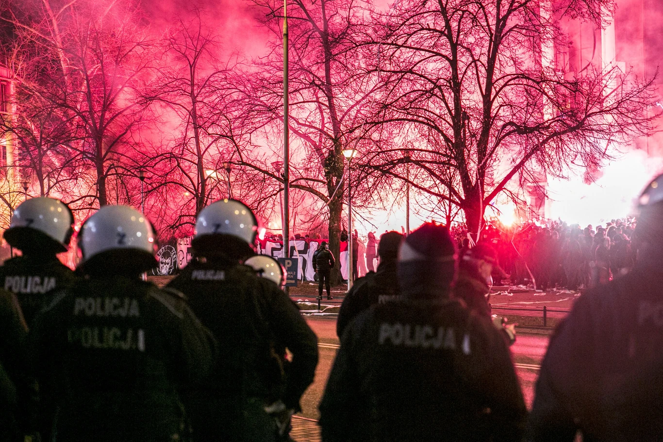 Grupa policjantów w kaskach i mundurach stoi plecami do obiektywu, zwrócona w stronę tłumu uczestników demonstracji, z których wielu trzyma race, generując intensywne czerwone światło i dym wśród gołych drzew na miejskiej ulicy.