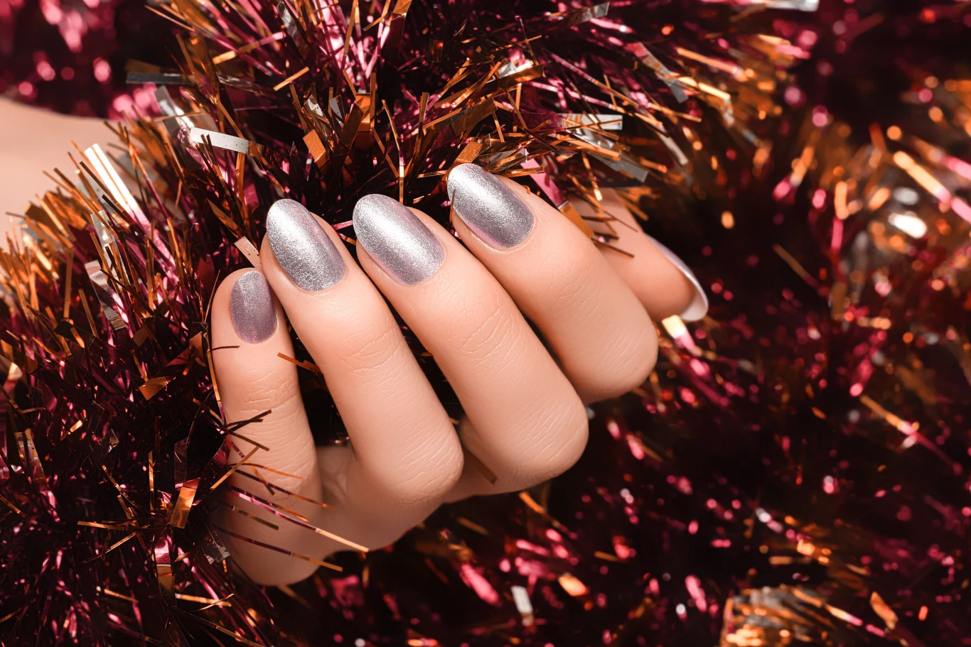 Dłoń z zadbanymi paznokciami pomalowanymi na metaliczny srebrny kolor wśród połyskujących fioletowo-złotych ozdób choinkowych.