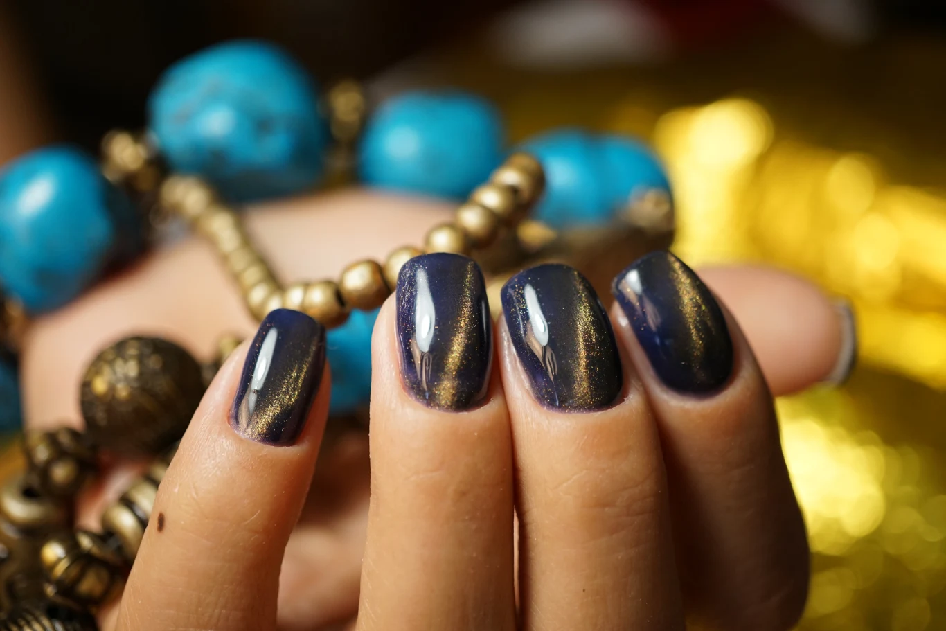 Dłoń z manicurem w odcieniu granatu i złota trzymająca niebieskie oraz złote koraliki, na drugim planie rozmyte tło o ciepłych barwach.