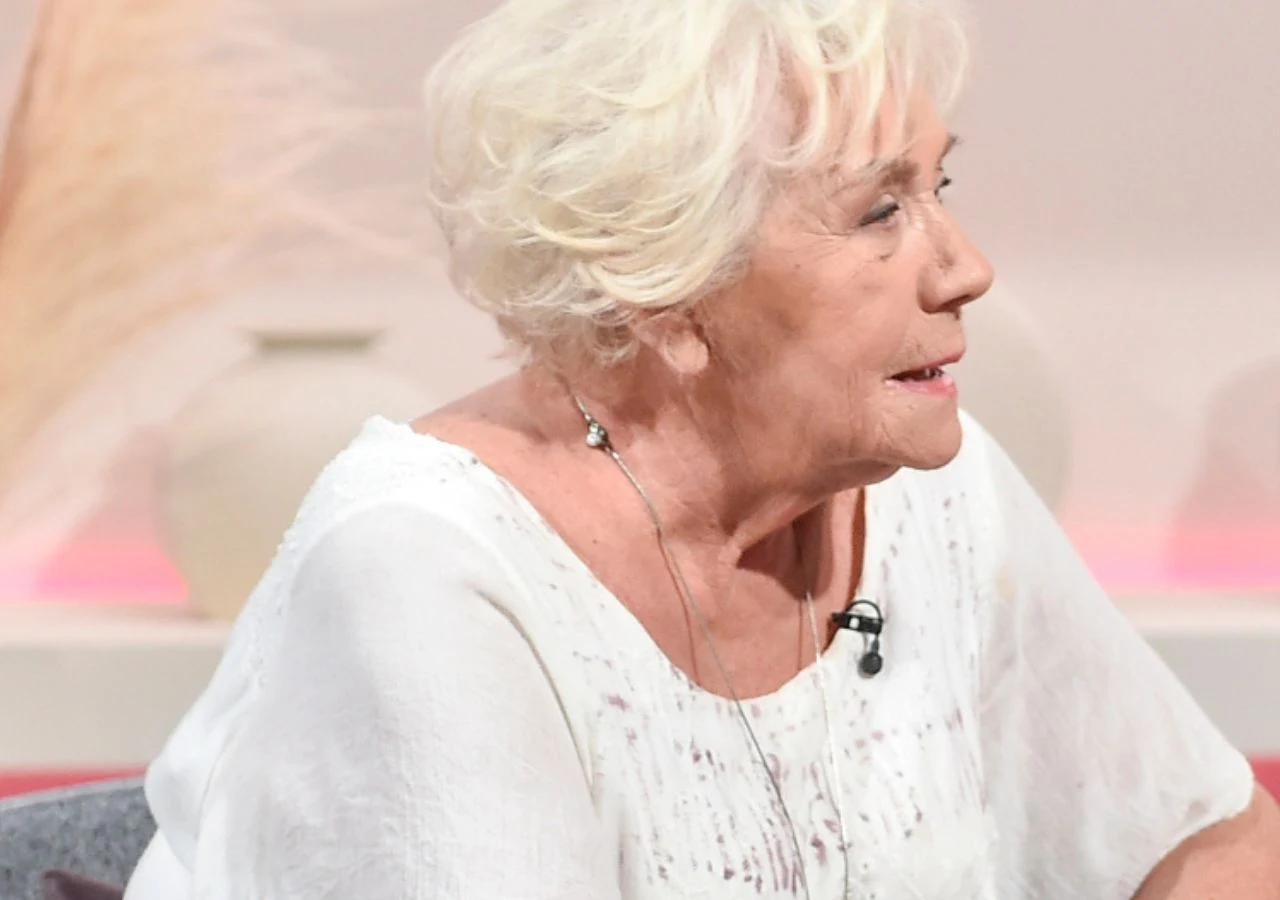 Teresa Lipowska Starsza kobieta z krótkimi, siwymi włosami siedzi w profilu na jasno oświetlonym tle, ubrana w białą bluzkę, z mikrofonem przypiętym przy dekolcie. W tle widoczne rozmyte elementy dekoracyjne.