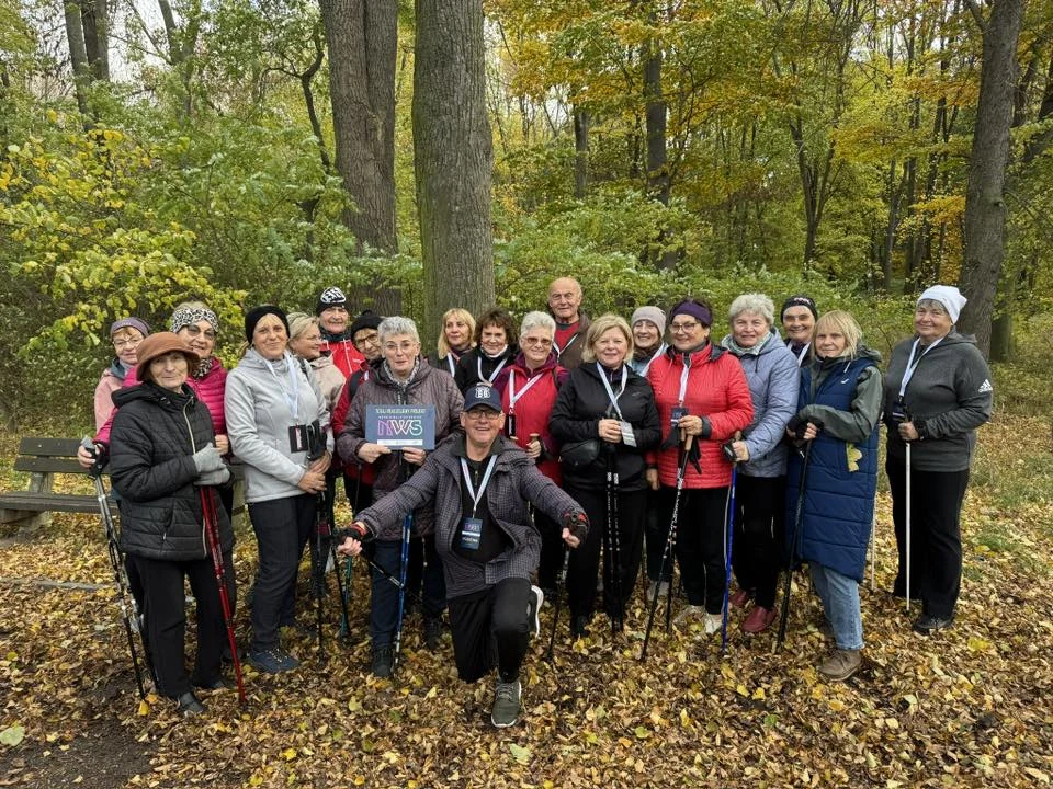 Na mapie Polski pojawiła się inicjatywa, która daje każdemu seniorowi szansę na udział w darmowych zajęciach ruchowych Grupa uśmiechniętych osób ubranych w sportowe kurtki i czapki, trzymających kijki do nordic walking, pozujących razem na tle jesiennego lasu wśród opadłych liści.