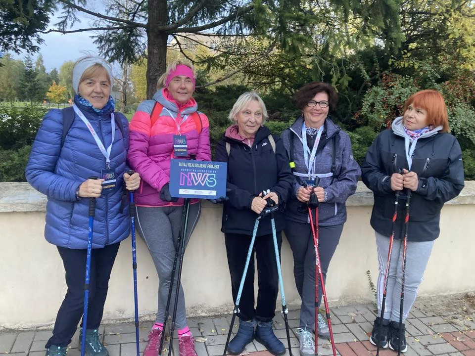 Regularne uprawianie nordic walkingu pozytywnie wpływa nie tylko na ciało, ale i umysł Pięć kobiet w sportowych ubraniach trzyma kijki do nordic walkingu i pozują razem podczas spaceru. Uśmiechnięte panie stoją na tle zielonych drzew i cementowego murku. Jedna z nich trzyma tabliczkę z napisem NWS.