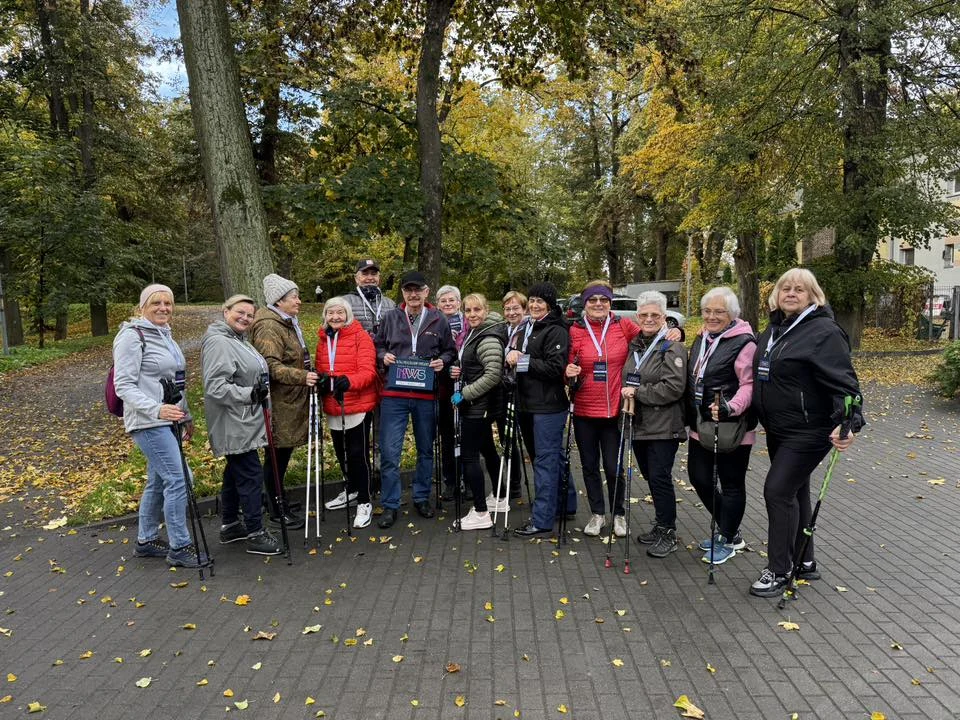 Nordic walking to coś pomiędzy joggingiem i szybkim biegiem. Największą różnicę robi jednak odciążenie stawów i zaangażowanie do 90 proc. mięśni Grupa starszych osób ubranych w jesienne kurtki pozuje razem na parkowej alejce trzymając kijki do nordic walking, w tle drzewa z jesiennymi liśćmi i fragmenty budynków