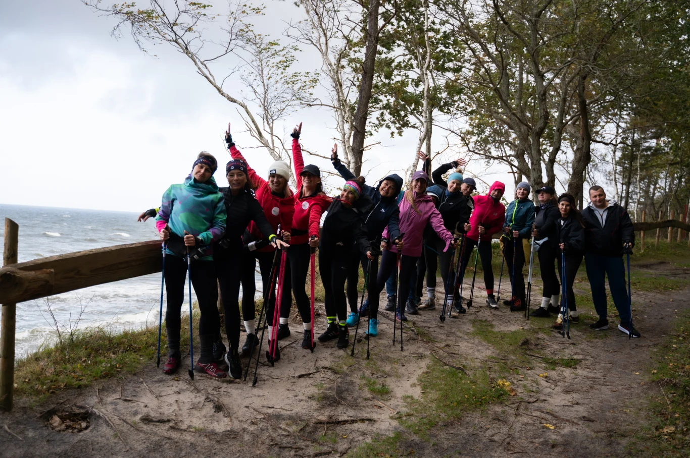 Polskie Stowarzyszenie Nordic Walking od lat zrzesza ludzi z pasją – niezależnie od wieku Grupa osób ubranych w sportowe stroje z kijkami do nordic walkingu pozuje wspólnie na leśnej ścieżce przy wybrzeżu, otoczona drzewami i widokiem na morze, w wietrzny i pochmurny dzień.