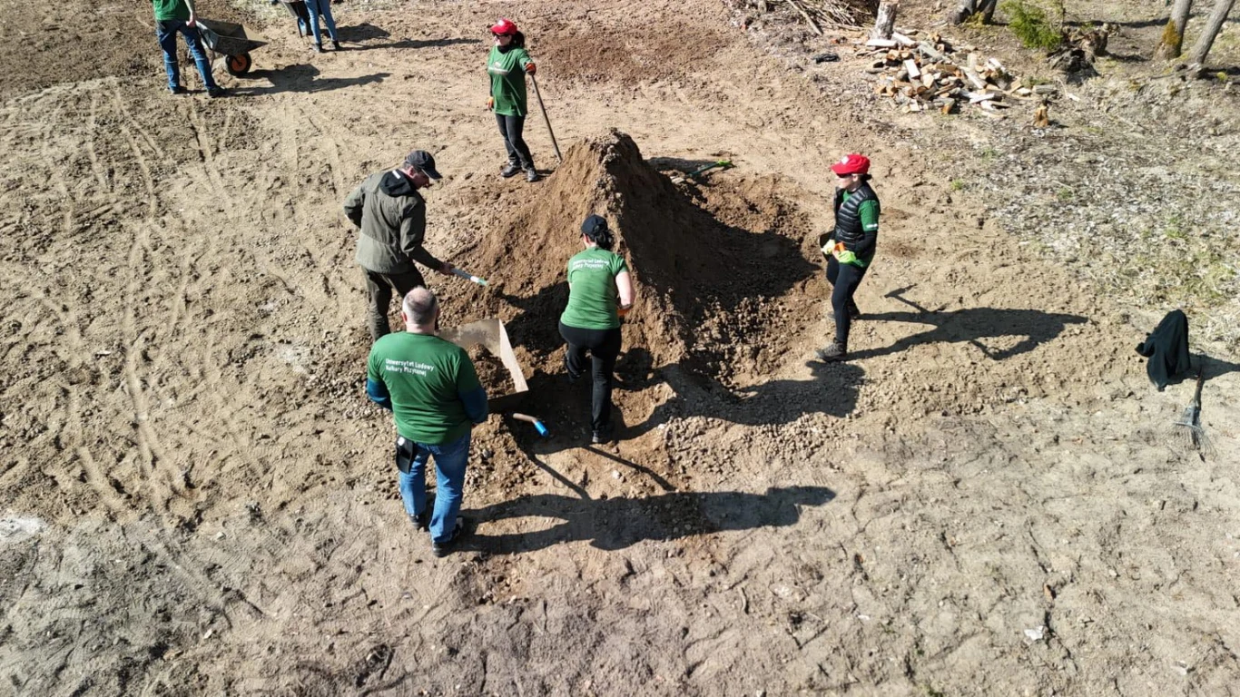 W PSNW wolontariat to połączenie działania, wspólnoty i realnych zmian, które często zaczynają się niepozornie Grupa osób w zielonych koszulkach i czerwonych czapkach pracuje wspólnie przy usypywaniu lub przerzucaniu stosu ziemi na otwartej przestrzeni, każda z nich używa łopaty, wokół widoczny teren leśny.