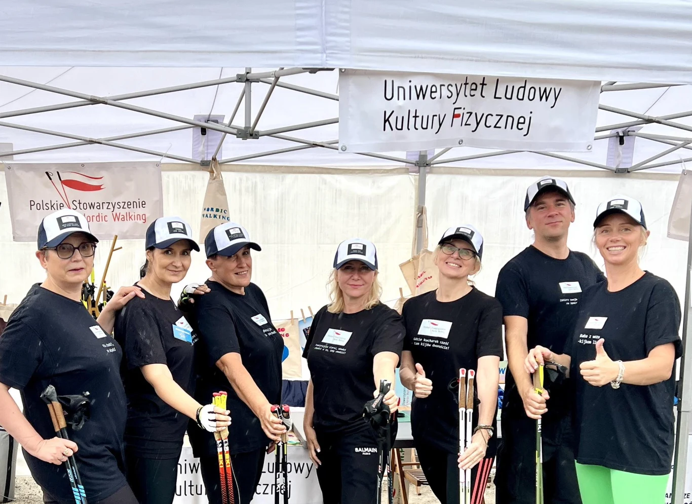 Dla wielu osób wolontariat w PSNW zaczyna się od zwykłej ciekawości Grupa siedmiu uśmiechniętych osób ubranych w czarne koszulki i czapki prezentuje kijki do nordic walkingu, stojąc pod namiotem z napisem Uniwersytet Ludowy Kultury Fizycznej i banerem Polskiego Stowarzyszenia Nordic Walking.