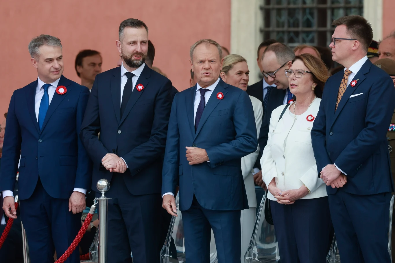 Przedstawiciele koalicji rządzącej od lewej: Krzysztof Gawkowski, wicepremier z Lewicy, Władysław Kosiniak-Kamysz, wicepremier z PSL, premier Donald Tusk, Małgorzata Kidawa-Błońska, marszałek Senatu z KO oraz Szymon Hołownia, były marszałek Sejmu z PL2050 Grupa elegancko ubranych osób w garniturach i marynarkach z przypiętymi biało-czerwonymi kotylionami zgromadzona podczas oficjalnej uroczystości, w tle ceglany mur i okno z kratą.