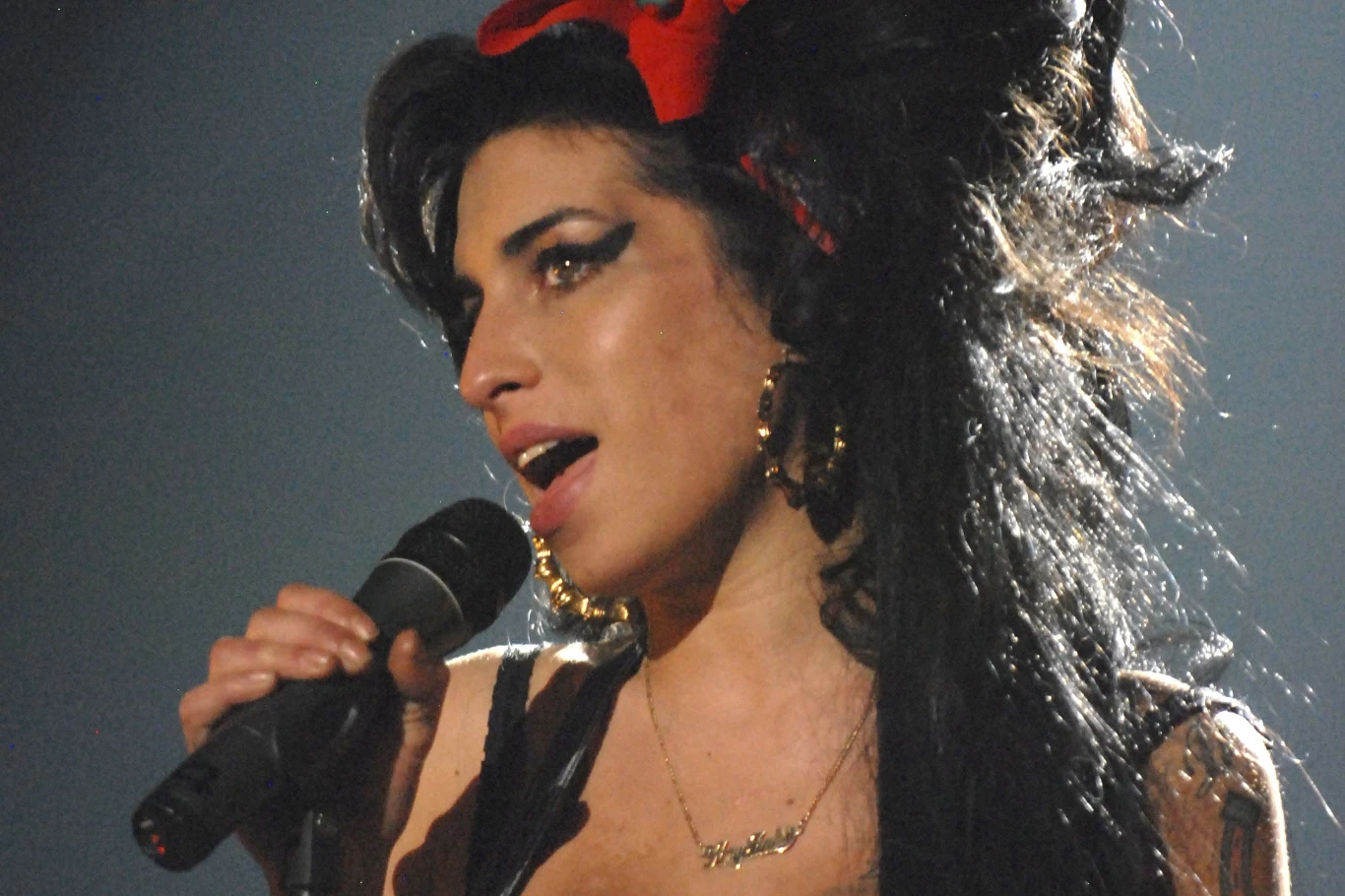 Rodzina i przyjaciółki Amy Winehouse spotkali się w sądzie Kobieta śpiewająca do mikrofonu na scenie, wyrazista charakteryzacja z mocnym makijażem oczu i charakterystyczną fryzurą, w uszach duże kolczyki, na głowie czerwona kokardka, intensywne oświetlenie podkreślające twarz artystki.