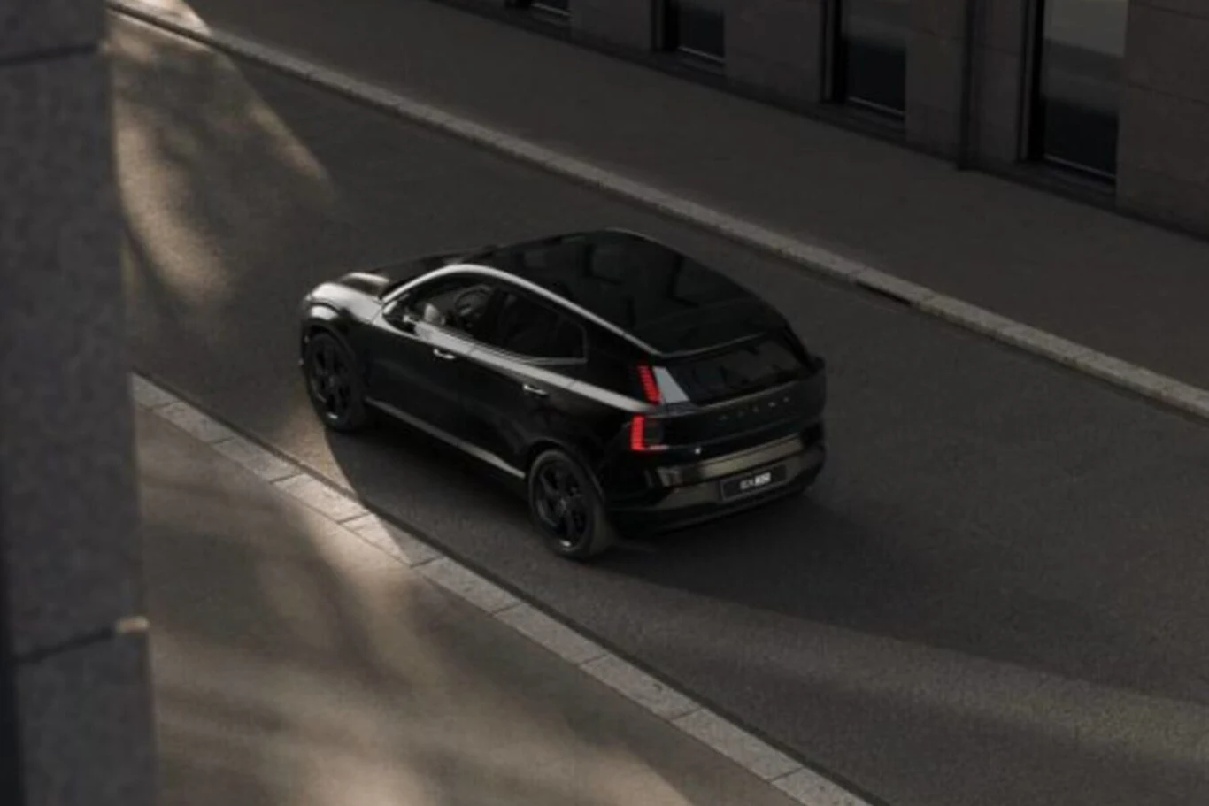 W standardzie Volvo EX30 znalazły się 19-calowe, czarne felgi Czarny nowoczesny samochód SUV jedzie pustą, asfaltową ulicą miejską w otoczeniu wysokich budynków z dużymi oknami.