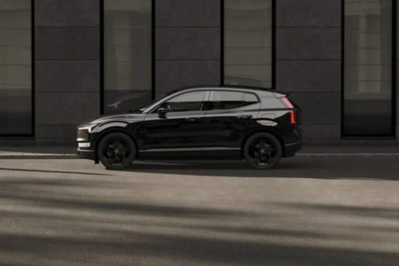 Volvo EX30 cechuje elegancka, jednolita stylistyka Czarny samochód typu SUV zaparkowany przy krawężniku na tle nowoczesnego budynku z dużymi, ciemnymi oknami.