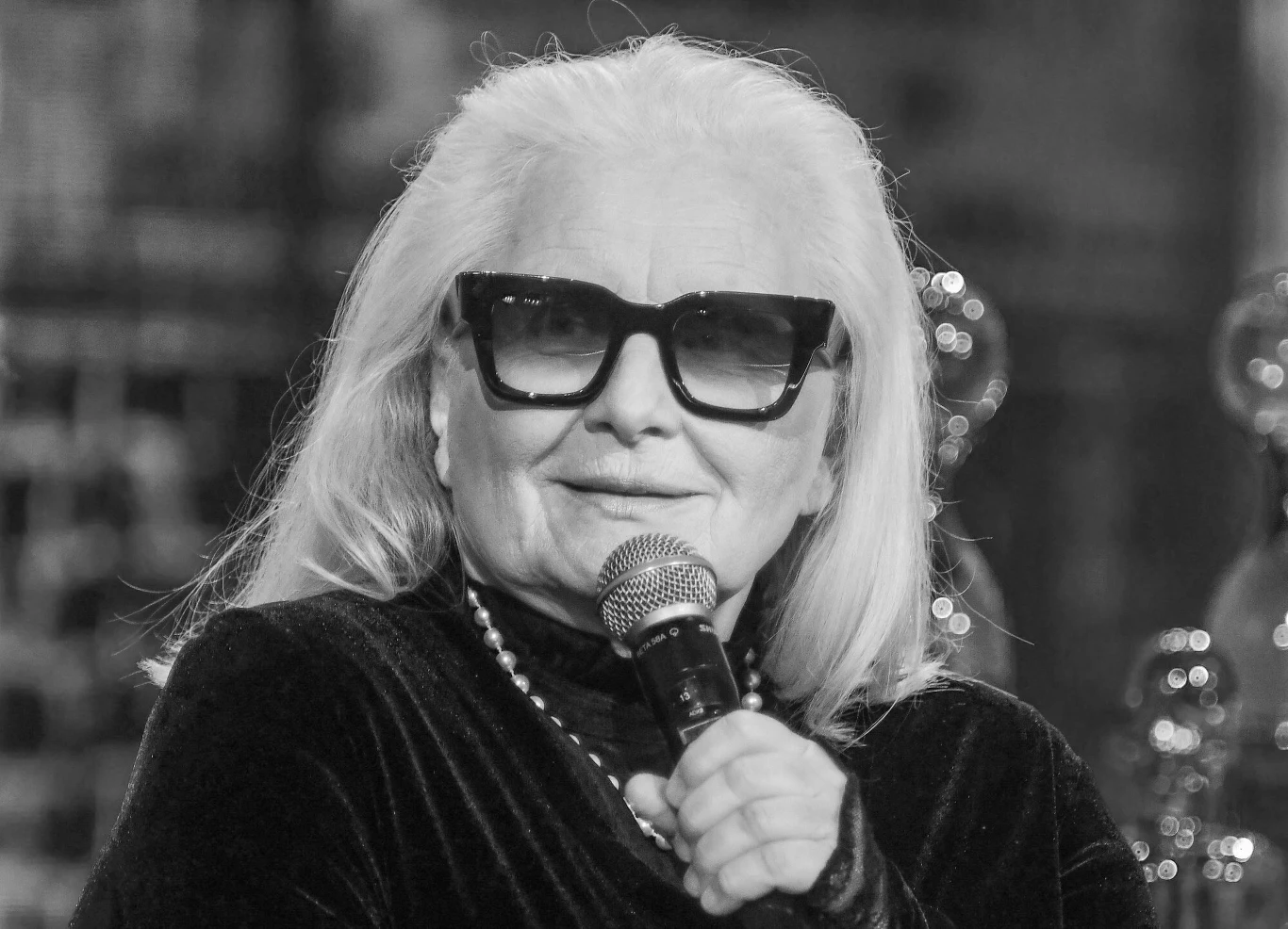 Magda Umer miała 76 lat Starsza kobieta w ciemnych okularach i eleganckim stroju, trzyma mikrofon i delikatnie się uśmiecha. Przewieszony przez szyję naszyjnik z pereł podkreśla jej szykowny wygląd.