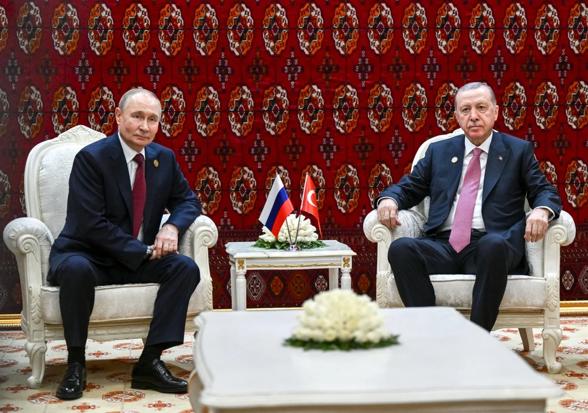 Putin i Erdogan rozmawiali w Turkmenistanie. Tematem wojna w Ukrainie Dwóch mężczyzn siedzących na białych fotelach przy stole, między nimi stoją flagi Rosji i Turcji ustawione na serwetce z kwiatami. Tło stanowi czerwony dekoracyjny dywan z orientalnym wzorem.