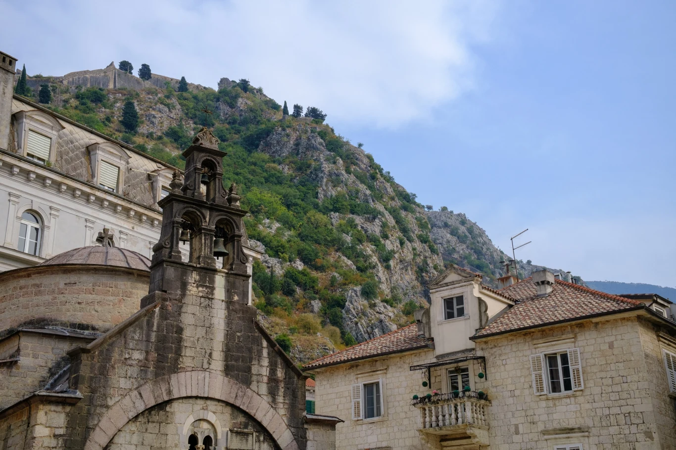Kamienna zabudowa z ozdobnym wejściem i dzwonnicą na pierwszym planie, otoczona innymi historycznymi budynkami, w tle zalesione zbocze góry pod błękitnym niebem.