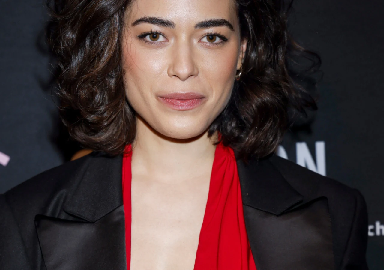 Młoda kobieta o krótkich, ciemnych włosach i jasnej cerze pozuje na tle ciemnej ścianki z nadrukiem. Ma na sobie elegancką, czarną marynarkę oraz czerwoną bluzkę z głębokim dekoltem, patrzy prosto w obiektyw i lekko się uśmiecha.