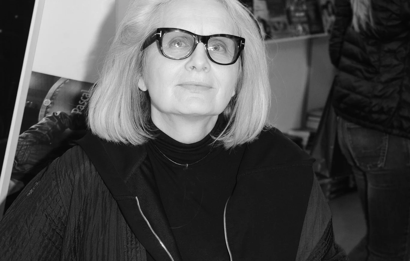 Starsza kobieta z jasnymi włosami i okularami siedzi w ciemnym ubraniu, z lekko uniesioną głową, patrząc przed siebie. W tle widać stojących ludzi i rozmyte półki z książkami.