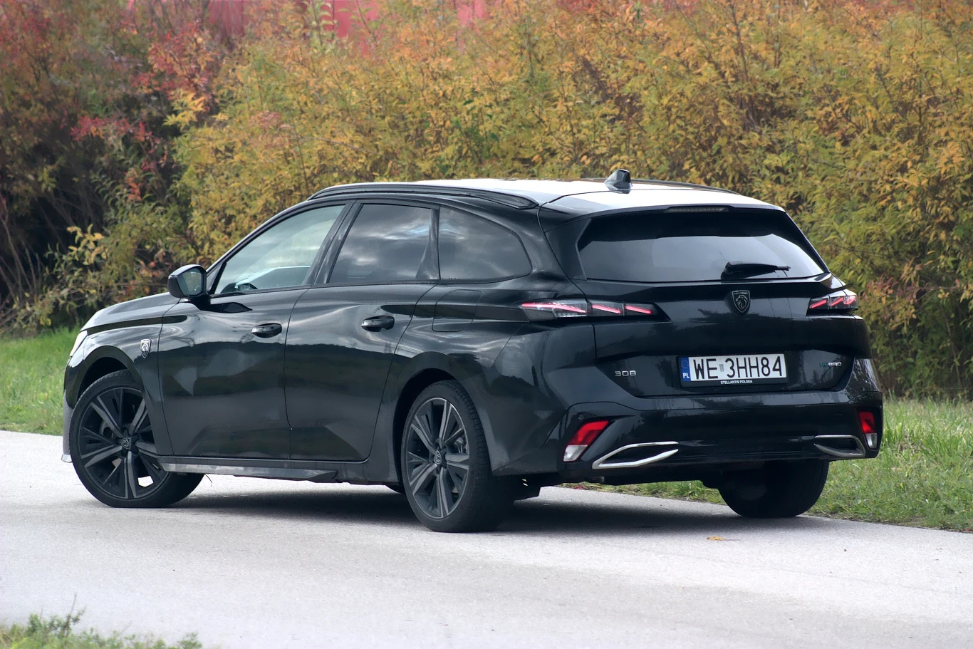 Czarny samochód osobowy typu kombi jadący wzdłuż asfaltowej drogi, otoczonej zielenią i krzewami w jesiennych barwach. Widoczne są tylne reflektory LED, wyraźna linia sylwetki oraz polska tablica rejestracyjna.