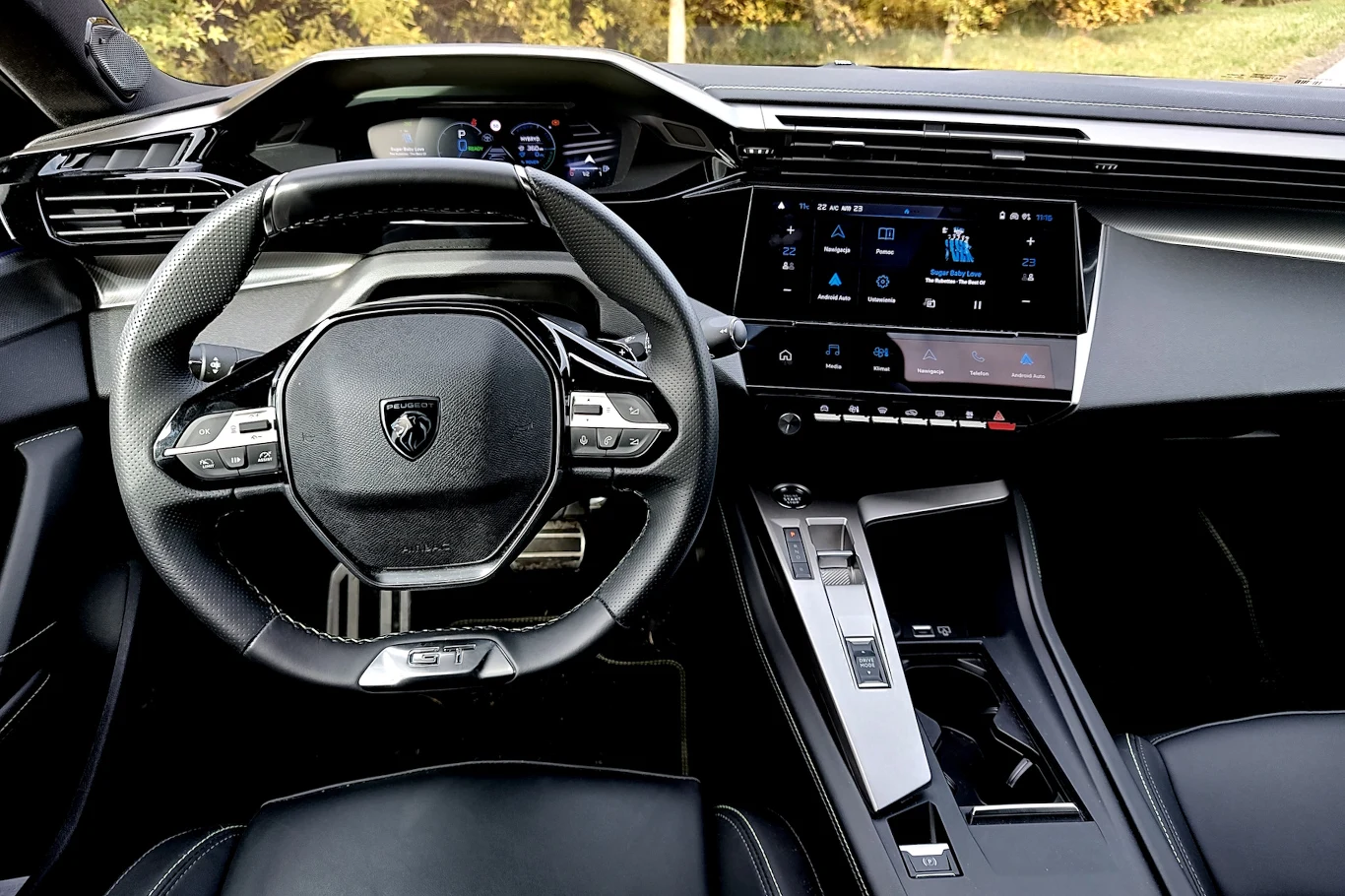 Kokpit nowoczesnego samochodu z kierownicą o sportowym wyglądzie, cyfrowymi wyświetlaczami, konsolą centralną z panelem dotykowym i przyciskami sterującymi funkcjami pojazdu.