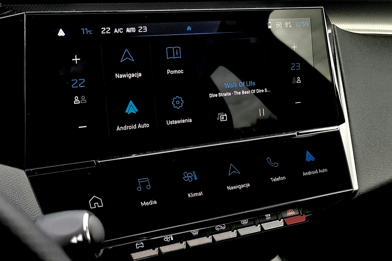 Dotykowy ekran samochodowego systemu multimedialnego z widocznymi ikonami nawigacji, pomocy, Android Auto, ustawień oraz aktualnie odtwarzaną muzyką. U dołu ekranu przyciski do sterowania mediami, klimatem, telefonem i innymi funkcjami.