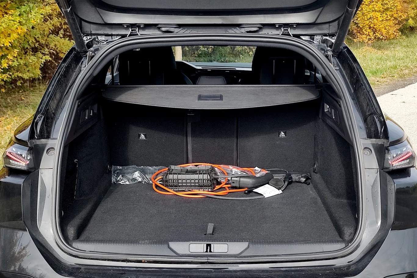 Otwarty bagażnik samochodu z widoczną ładowarką do pojazdów elektrycznych oraz pomarańczowym kablem ułożonym na czarnej wykładzinie, w tle jesienne krzewy i trawa.