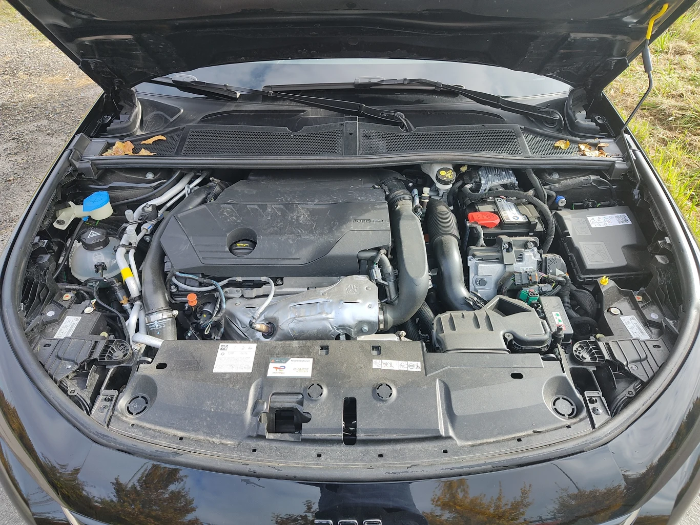 Otwarta maska samochodu odsłania silnik z widocznymi przewodami, akumulatorem, zbiornikiem płynu chłodniczego i innymi elementami technicznymi.